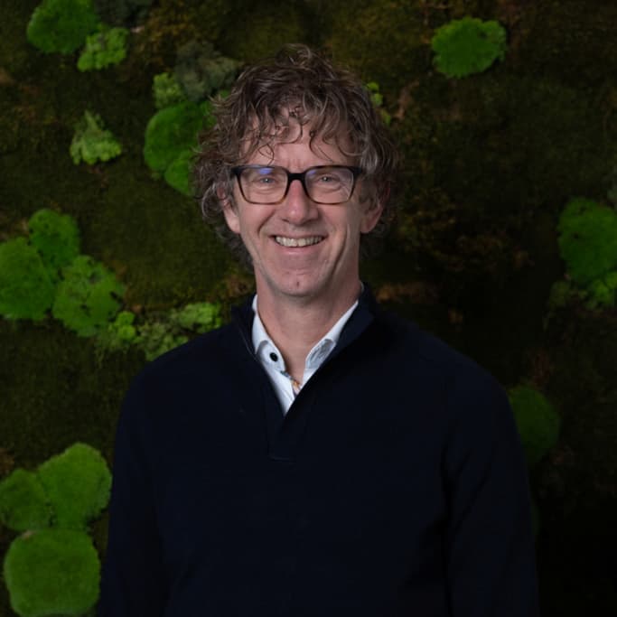 Rob Oud portret met bril in donkere trui tegen moswand